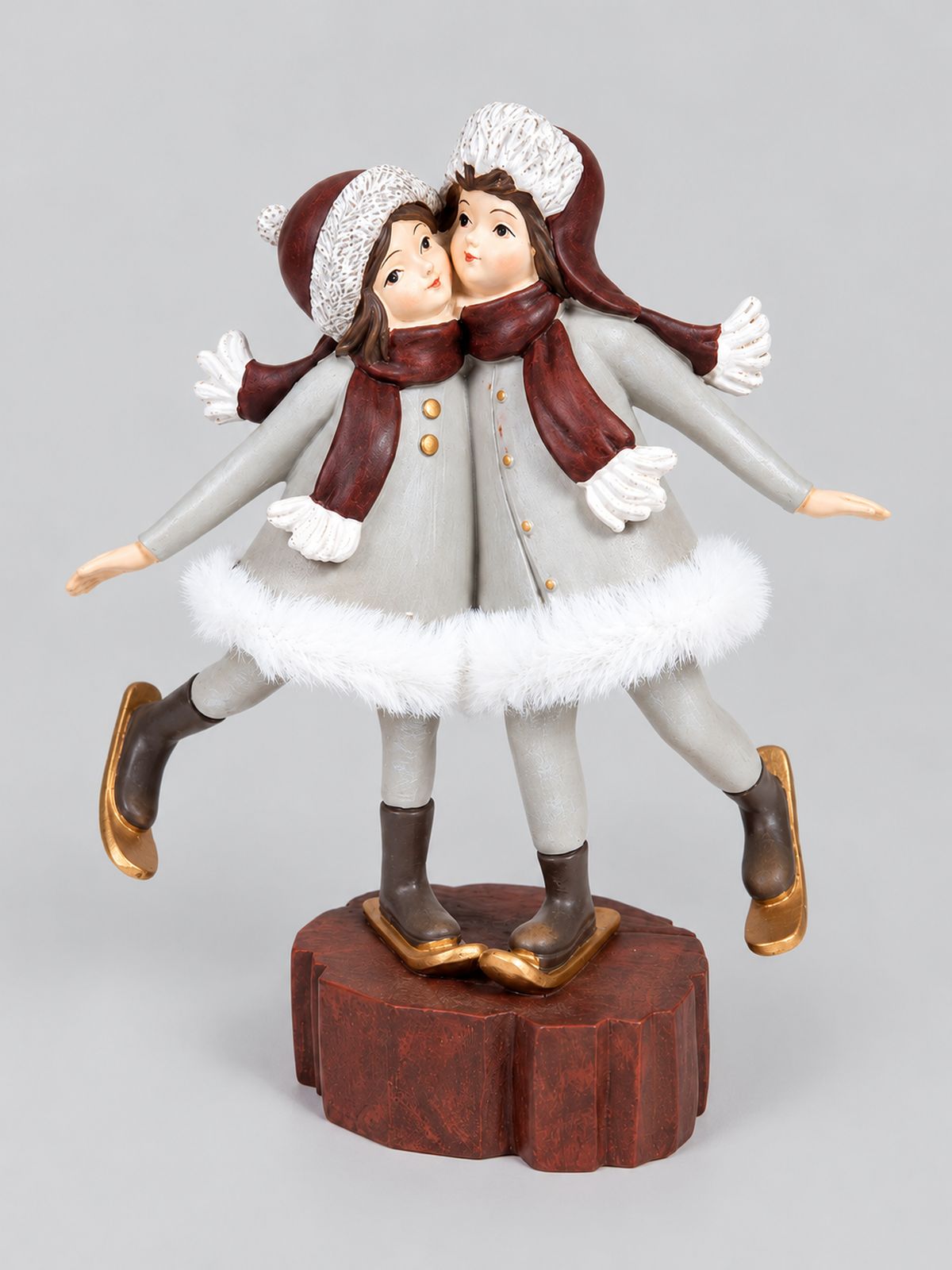 Figurine Les Petites Skieuses, deux enfants en manteaux gris et écharpes bordeaux sur un socle brun, présentée sur fond gris clair.