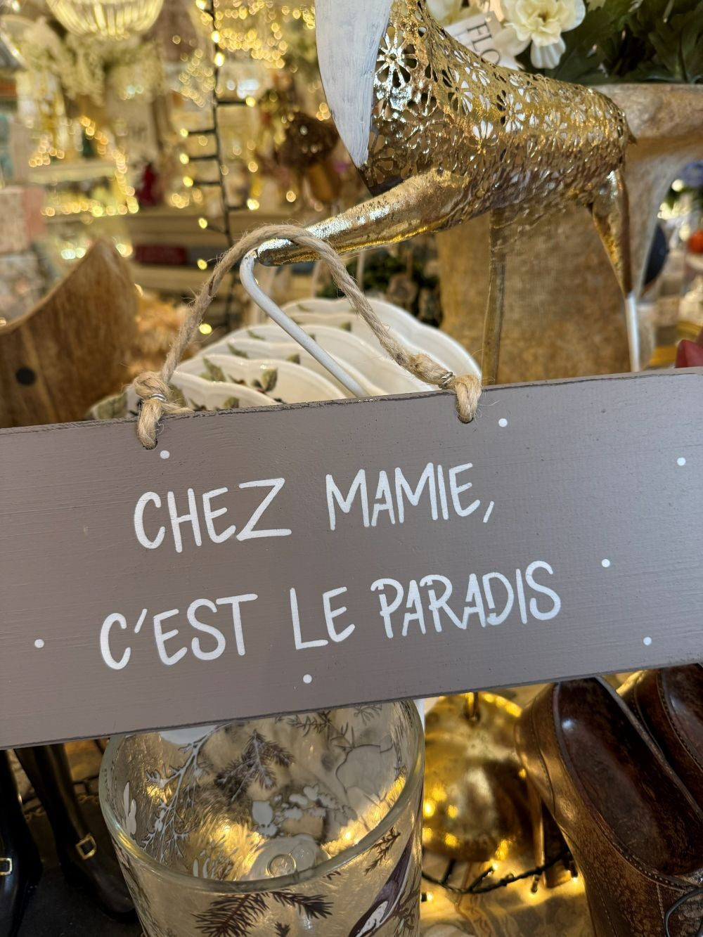 Plaque décorative en bois taupe avec inscription blanche « Chez Mamie c’est le paradis », style shabby chic, suspendue par une corde naturelle dans une ambiance romantique et chaleureuse