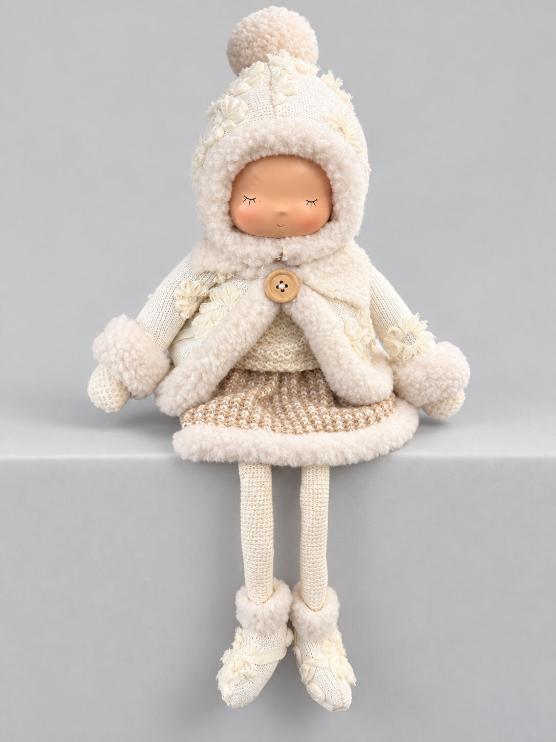 Grande poupée décorative d’hiver écrue et beige avec bonnet, tenue douillette et longues jambes, assise sur un meuble dans une ambiance shabby chic romantique