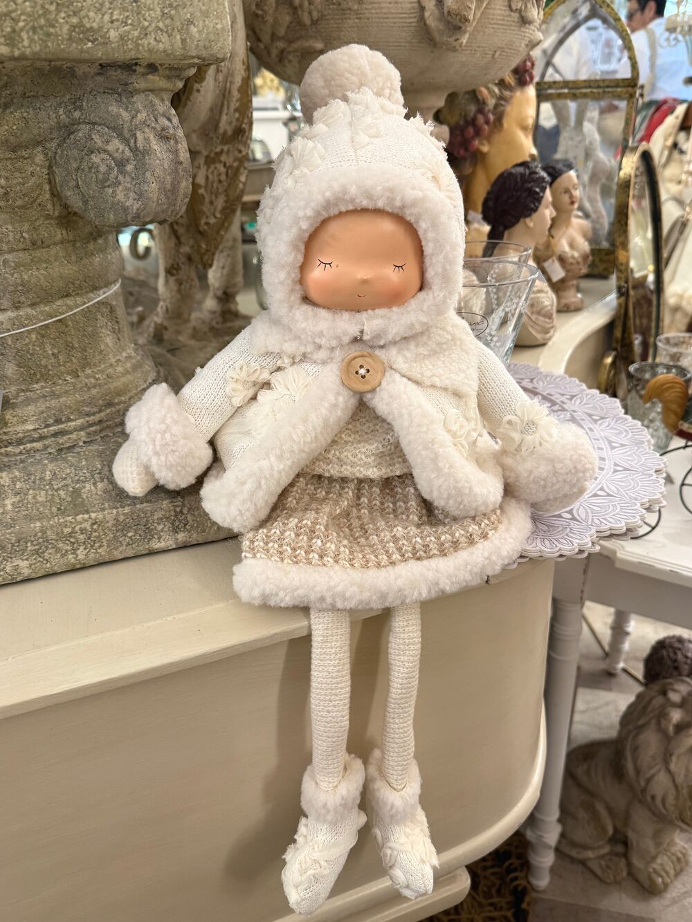 Grande poupée décorative d’hiver écrue et beige avec bonnet, tenue douillette et longues jambes, assise sur un meuble dans une ambiance shabby chic romantique