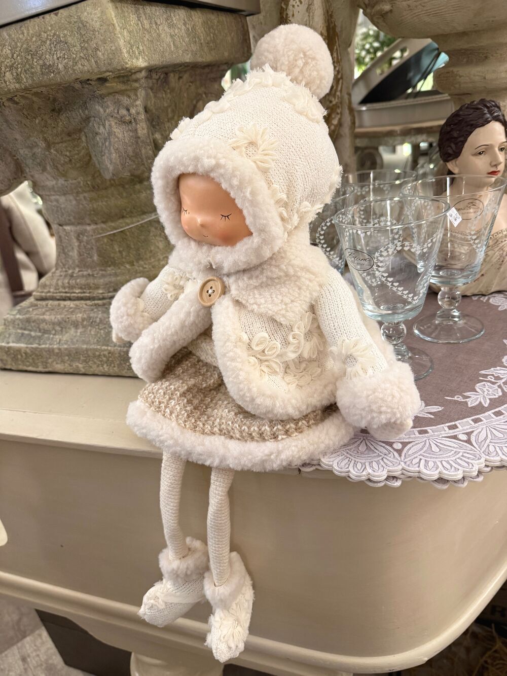 Grande poupée décorative d’hiver écrue et beige avec bonnet, tenue douillette et longues jambes, assise sur un meuble dans une ambiance shabby chic romantique
