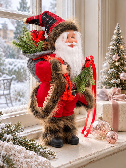 Poupée Père Noël Tradition près d’une fenêtre hivernale, accompagnée d’un cadeau et de boules roses.