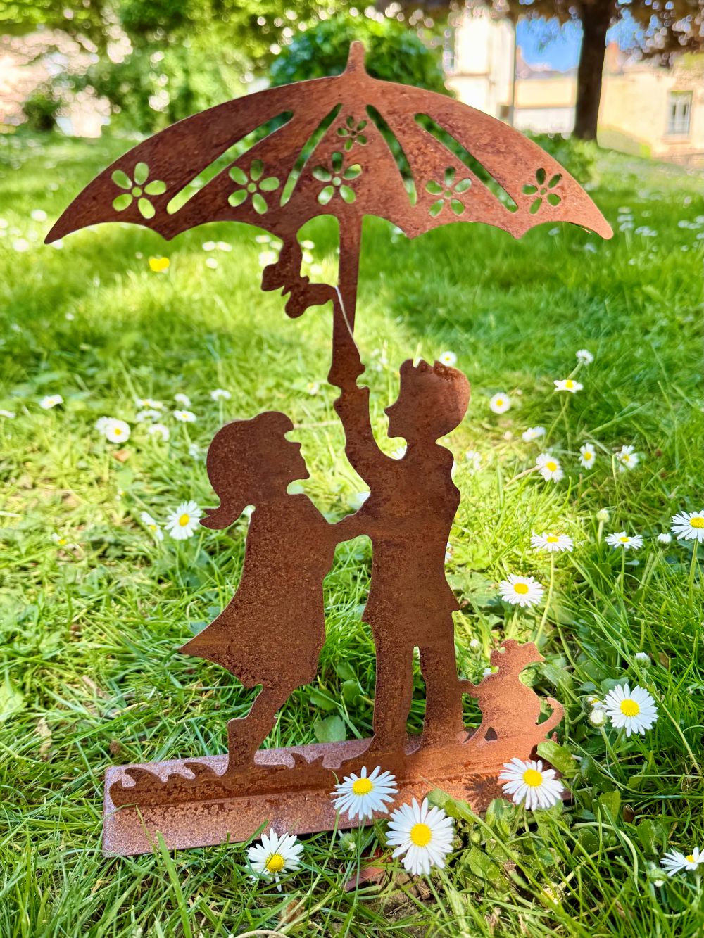 Silhouette décorative en fer forgé rouille représentant deux enfants sous une ombrelle avec un chien, posée sur une pelouse ensoleillée dans un jardin romantique.