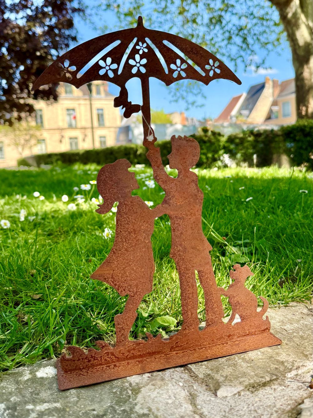 Silhouette décorative en fer forgé rouille représentant deux enfants sous une ombrelle avec un chien, posée sur une pelouse ensoleillée dans un jardin romantique.
