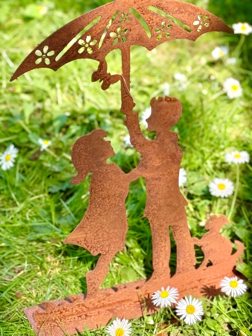Silhouette décorative en fer forgé rouille représentant deux enfants sous une ombrelle avec un chien, posée sur une pelouse ensoleillée dans un jardin romantique.