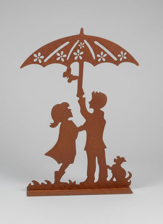 Silhouette décorative en fer forgé rouille représentant deux enfants sous une ombrelle avec un chien, posée sur une pelouse ensoleillée dans un jardin romantique.