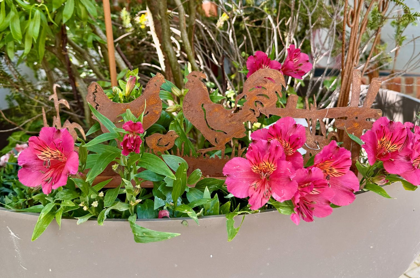 Silhouette décorative en fer forgé rouillé représentant une scène de basse-cour avec coq, poule et poussins, installée dans l’herbe d’un jardin champêtre