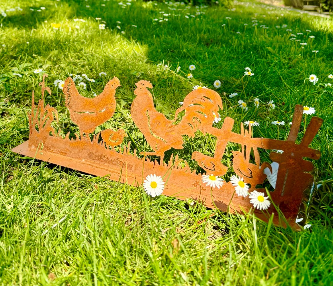 Silhouette décorative en fer forgé rouillé représentant une scène de basse-cour avec coq, poule et poussins, installée dans l’herbe d’un jardin champêtre