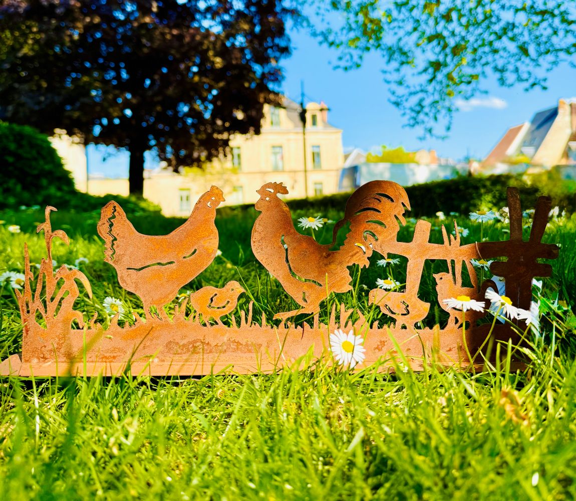 Silhouette décorative en fer forgé rouillé représentant une scène de basse-cour avec coq, poule et poussins, installée dans l’herbe d’un jardin champêtre