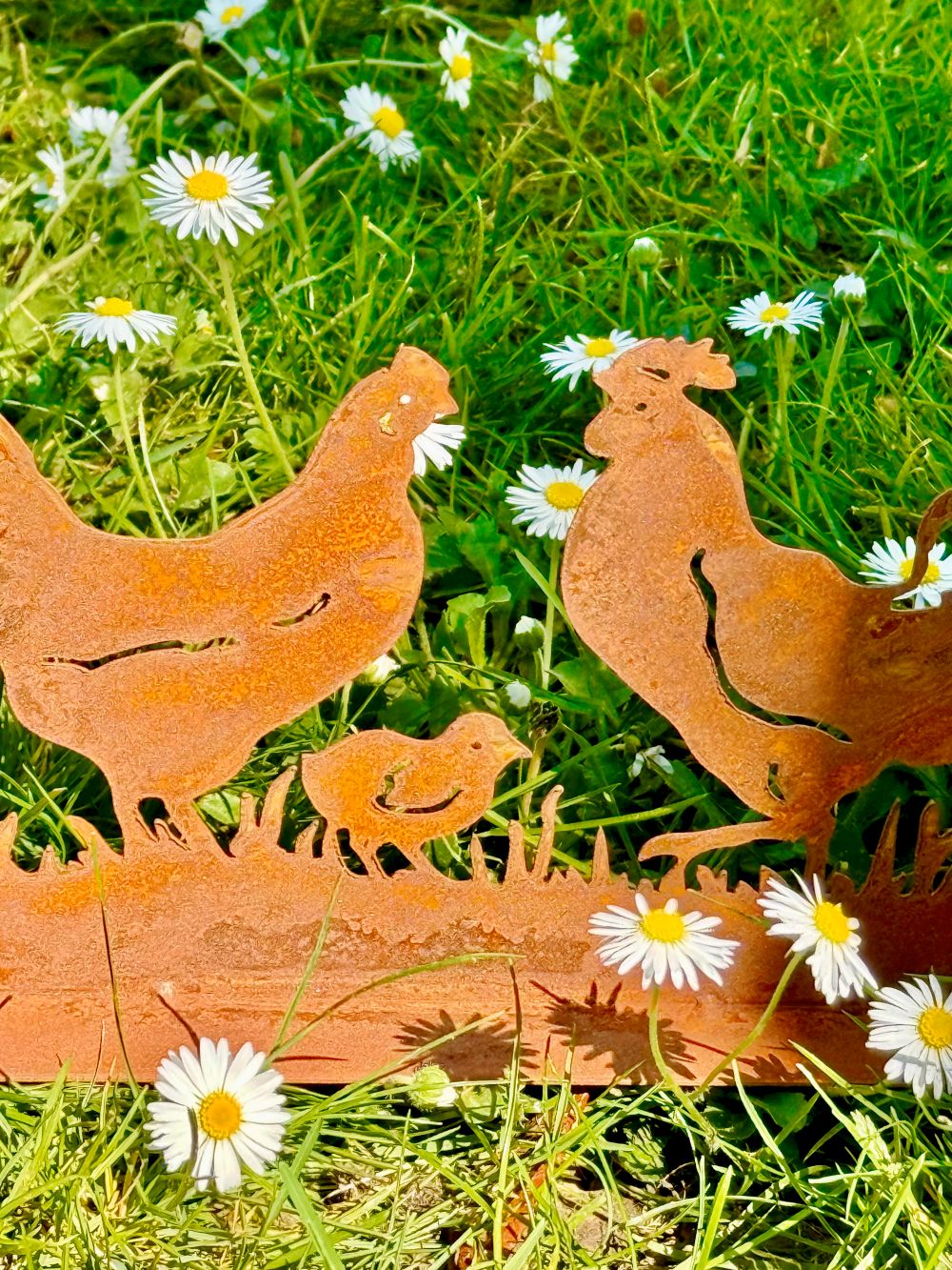 Silhouette décorative en fer forgé rouillé représentant une scène de basse-cour avec coq, poule et poussins, installée dans l’herbe d’un jardin champêtre