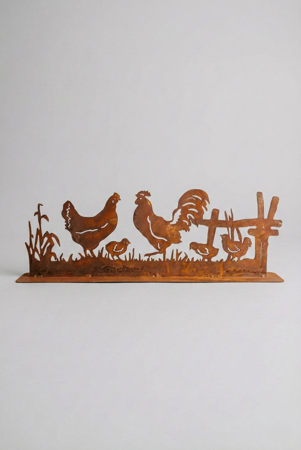 Silhouette décorative en fer forgé rouillé représentant une scène de basse-cour avec coq, poule et poussins, installée dans l’herbe d’un jardin champêtre