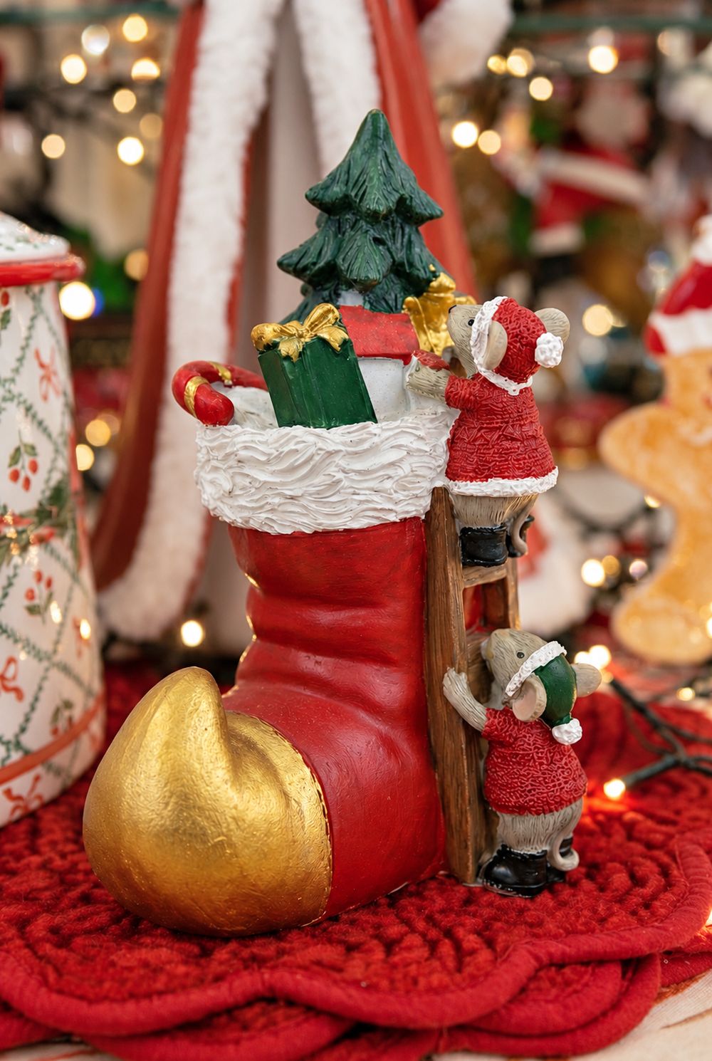 Soulier de Noël rouge et doré, avec sapin et deux souris sur une échelle, dans un décor de Noël aux lumières floues.