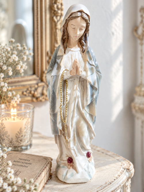 Statuette décorative de la Vierge Marie en prière tenant un chapelet doré, finitions nacrées blanc cassé, détails roses à la base, style romantique et shabby chic, ambiance spirituelle et poétique