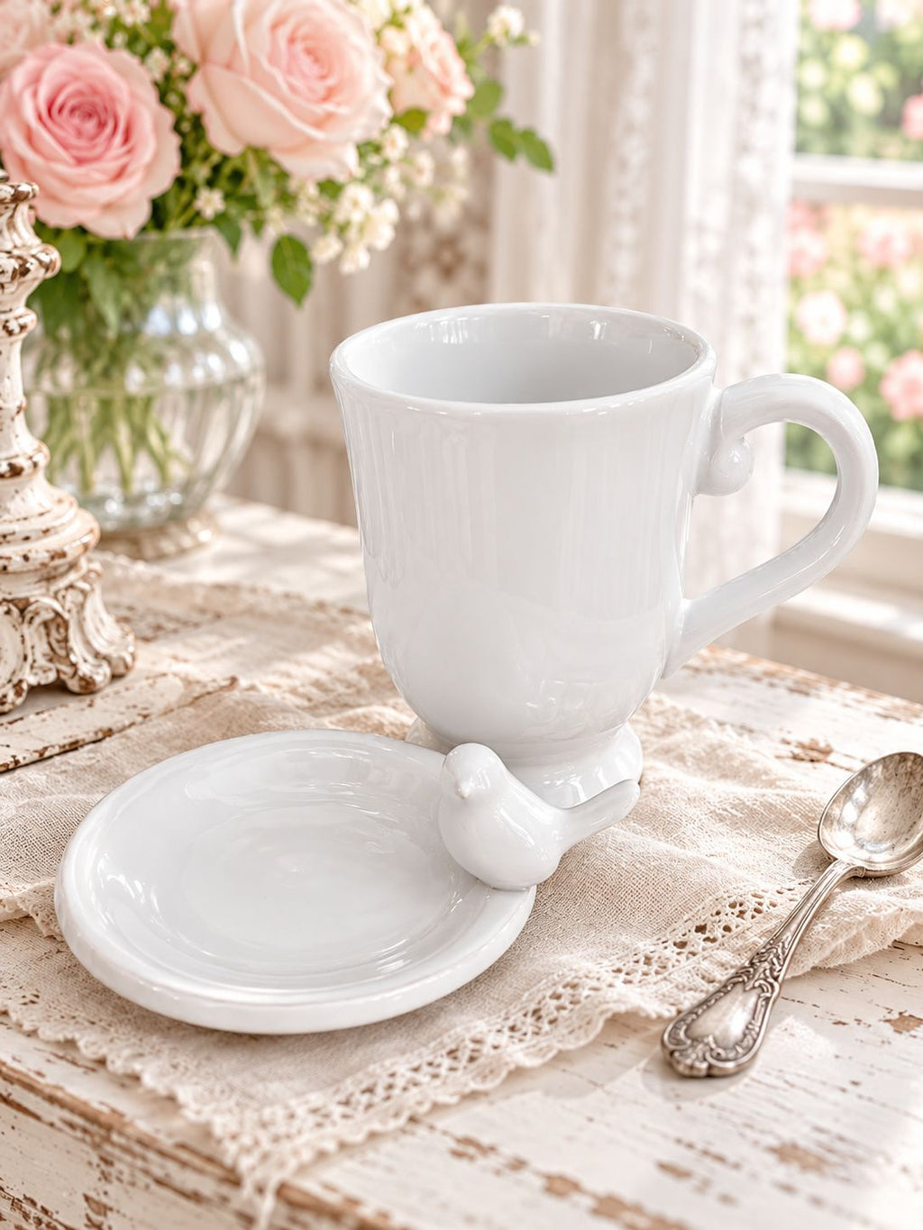 Tasse Chant de l’Oiseau et sa coupelle sur une table patinée, près de roses roses et d’une fenêtre.