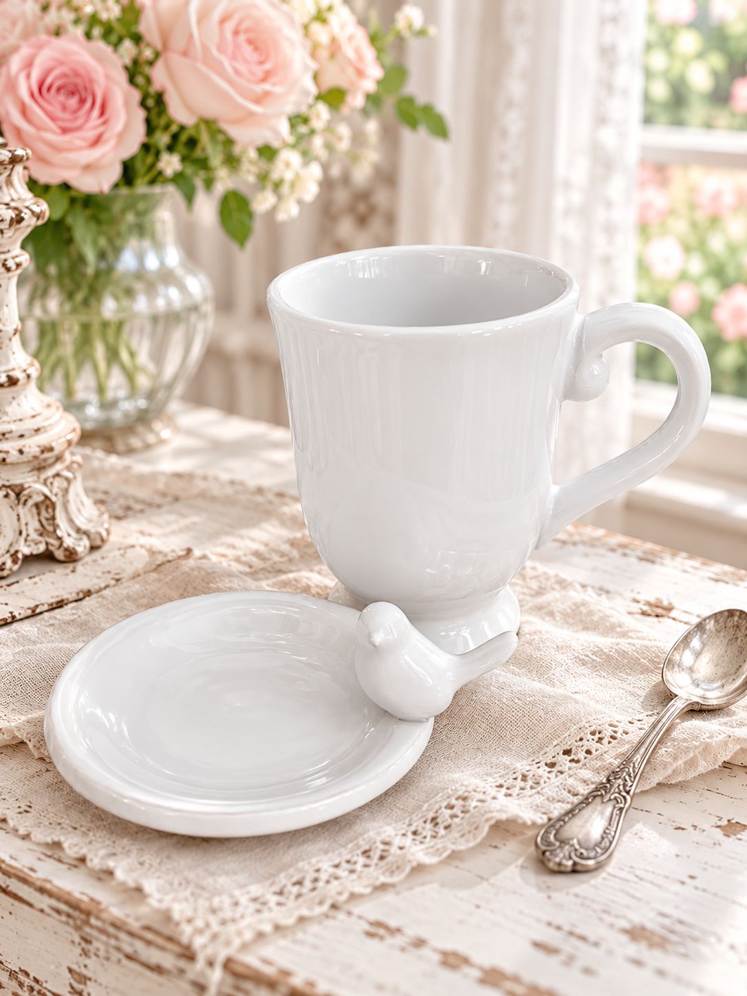 Tasse Chant de l’Oiseau et sa coupelle sur une table patinée, près de roses roses et d’une fenêtre.