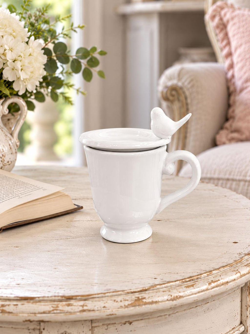 Tasse blanche fermée par sa coupelle oiseau, sur une table près d’un livre et d’un fauteuil.