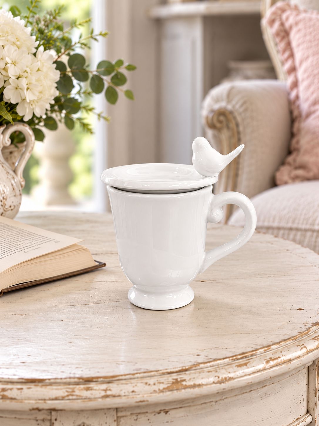 Tasse blanche fermée par sa coupelle oiseau, sur une table près d’un livre et d’un fauteuil.