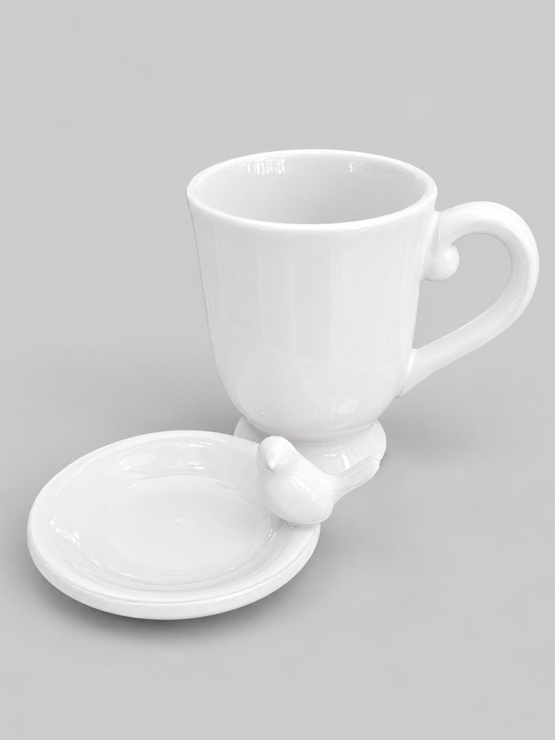 Tasse blanche en faïence et coupelle ornée d’un oiseau, présentées sur fond gris clair.