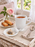 Tasse de thé fumant et coupelle oiseau accueillant une mousseline, sur un plateau shabby chic avec financiers et rose près d’une fenêtre.