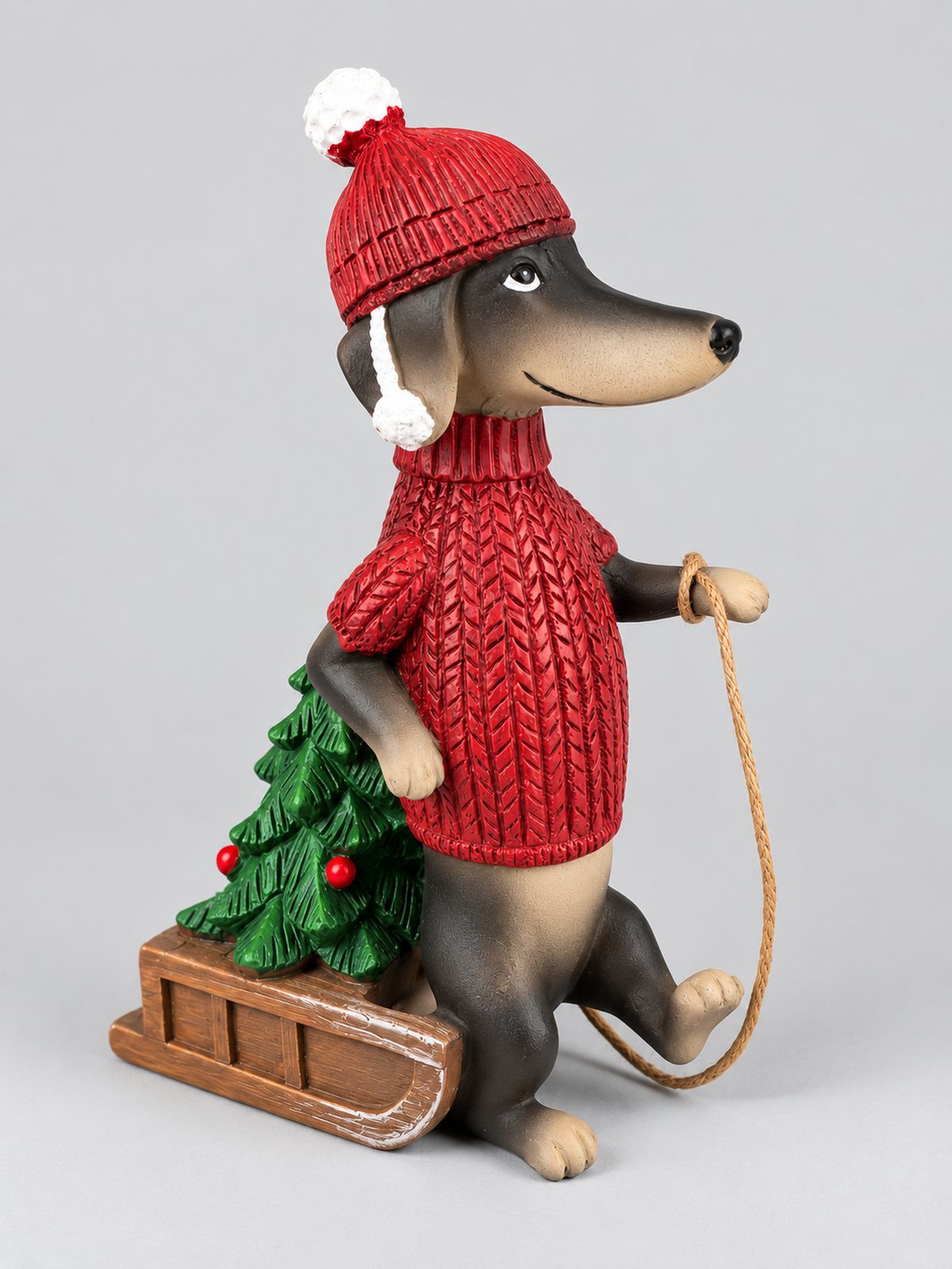 Figurine Teckel des Neiges en pull et bonnet rouges, avec une luge portant un sapin, sur fond gris clair.
