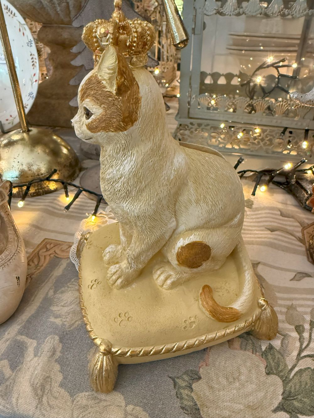 Tirelire décorative en forme de chat couronné, couleurs beige, doré et marron, style shabby chic romantique et poétique, posée sur un coussin élégant